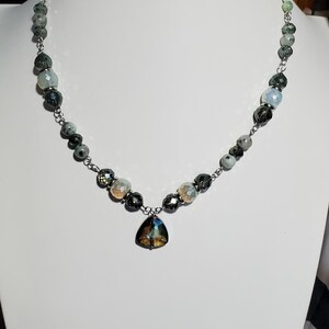 May include: A silver chain necklace with a teardrop-shaped pendant. The necklace is made with faceted beads in shades of green, gray, and white.