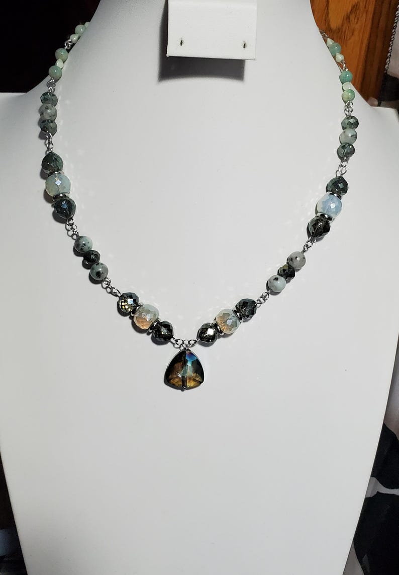 May include: A silver chain necklace with a teardrop-shaped pendant. The necklace is made with faceted beads in shades of green, gray, and white.