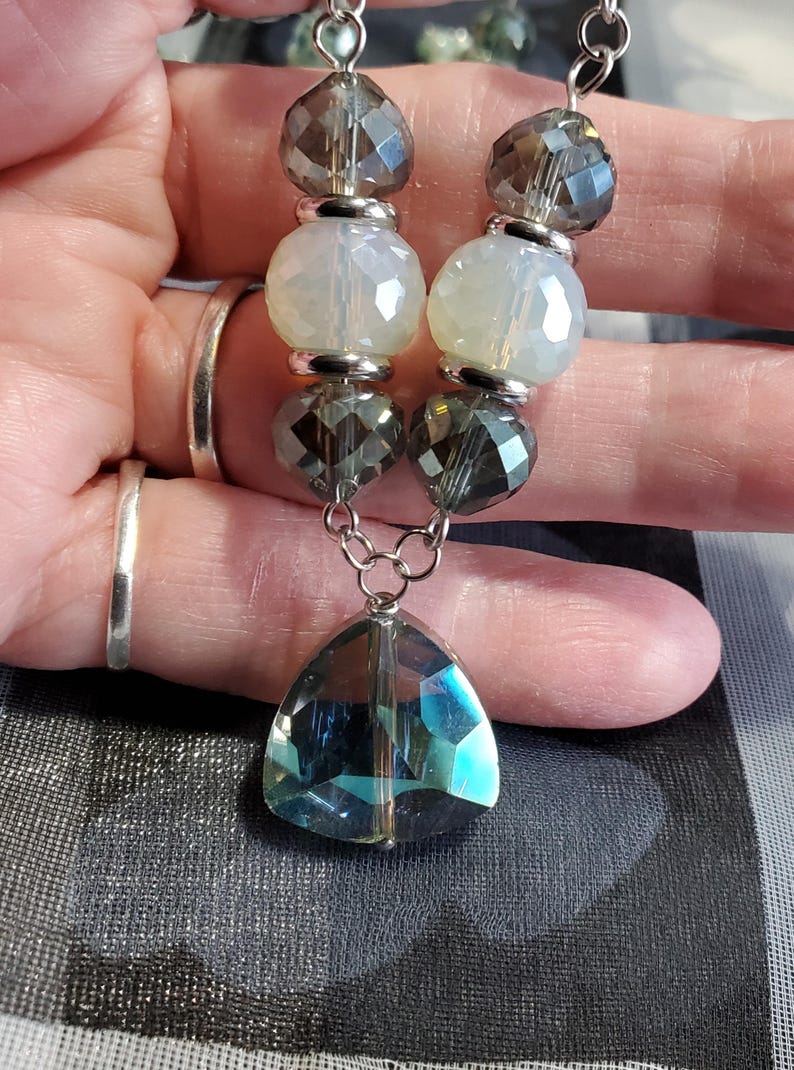 May include: A silver chain necklace with alternating clear, iridescent, and dark gray beads. The necklace has a large, teardrop-shaped, iridescent crystal pendant.