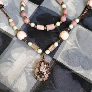 May include: A delicate necklace with a pink and green stone pendant. The pendant is set in a copper frame with floral designs. The necklace is made with pink and green beads and a copper chain.