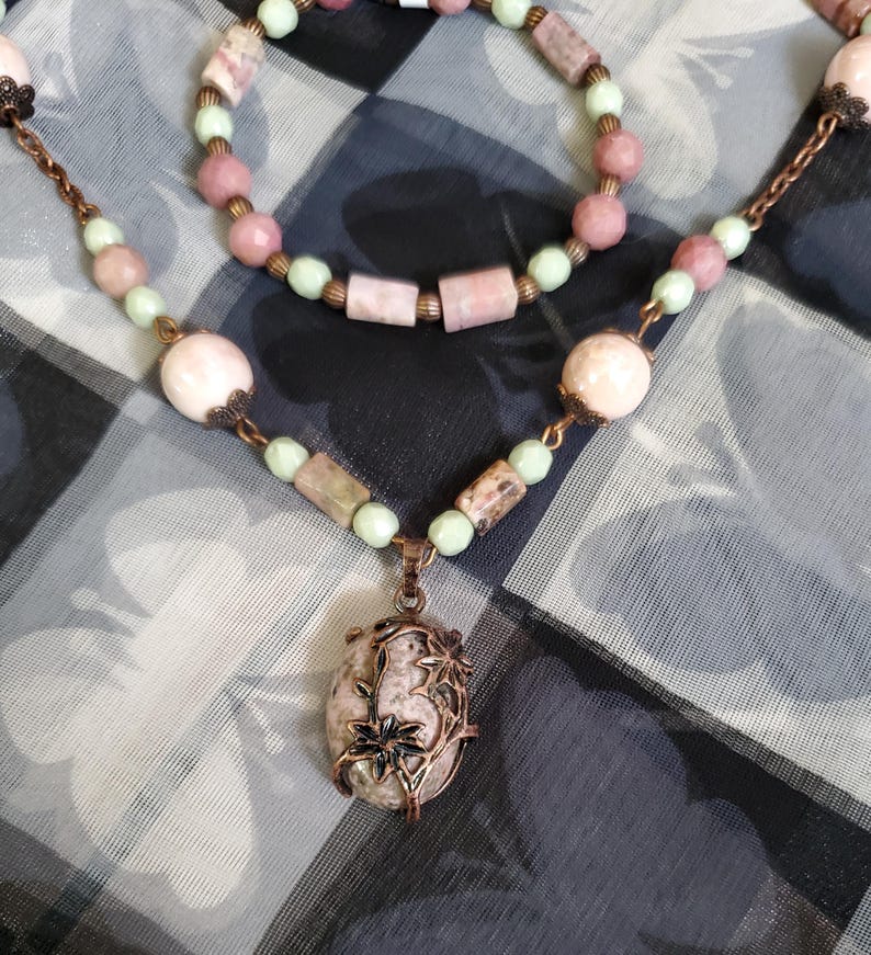 May include: A delicate necklace with a pink and green stone pendant. The pendant is set in a copper frame with floral designs. The necklace is made with pink and green beads and a copper chain.