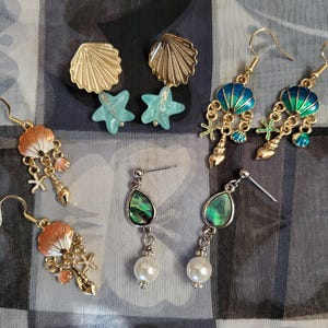 May include: A set of six pairs of earrings featuring a variety of sea-themed designs. The earrings are made of gold-tone metal and feature seashells, starfish, and pearls.