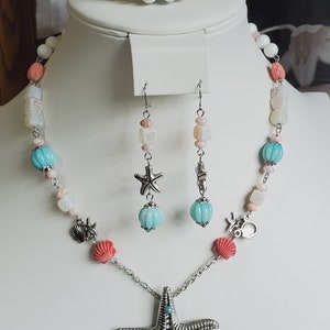 May include: A set of sea-themed jewellery featuring a necklace, earrings, and a bracelet. The necklace has a large silver starfish pendant, white and pink beads, and turquoise blue beads. The earrings are silver with turquoise blue beads and a starfish charm. The bracelet is white with turquoise blue beads and a starfish charm.