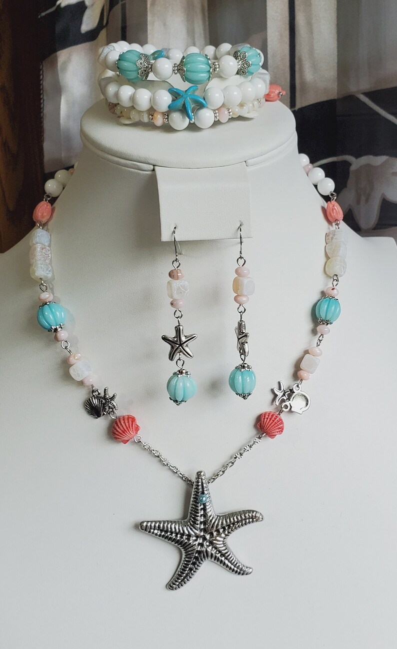 May include: A set of sea-themed jewellery featuring a necklace, earrings, and a bracelet. The necklace has a large silver starfish pendant, white and pink beads, and turquoise blue beads. The earrings are silver with turquoise blue beads and a starfish charm. The bracelet is white with turquoise blue beads and a starfish charm.
