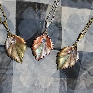 May include: Three leaf-shaped pendants with iridescent stones hanging from chains. The pendants are made of copper, brass, and silver, and each has a different color scheme.