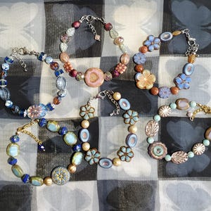 May include: Six beaded bracelets with various designs and colors are displayed. The bracelets feature a mix of blue, gold, pink, and other colorful beads. Some bracelets have floral and leaf-shaped accents, while others have unique geometric patterns.