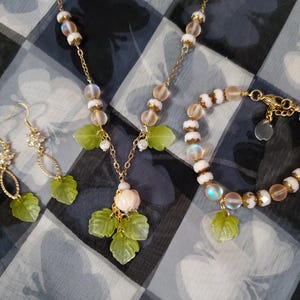 May include: A set of delicate jewelry featuring a necklace, bracelet, and earrings. The pieces are made with white and iridescent beads and have green leaf charms. The necklace has a small pink flower charm.