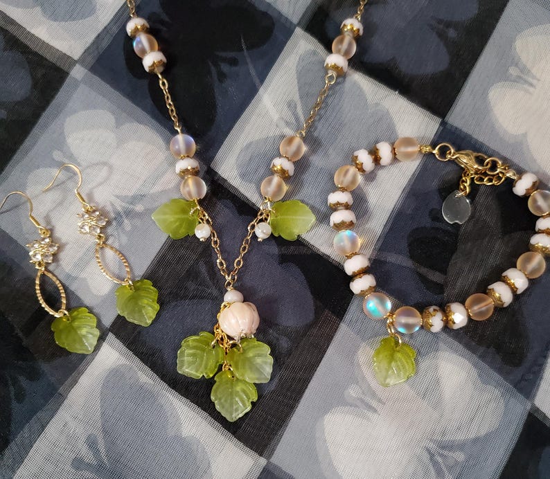 May include: A set of delicate jewelry featuring a necklace, bracelet, and earrings. The pieces are made with white and iridescent beads and have green leaf charms. The necklace has a small pink flower charm.
