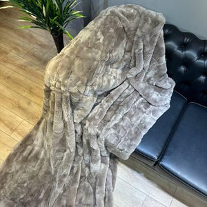 May include: A plush, faux fur blanket in a neutral taupe color. The blanket is draped over a black leather couch, showcasing its soft texture and rectangular pattern. The blanket's dimensions are not visible.