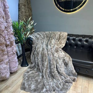 May include: A plush, gray faux fur throw blanket draped over a black leather sofa. The blanket has a soft, fluffy texture and is perfect for adding warmth and comfort to any room.