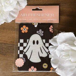 May include: Air freshener featuring a white ghost design on a black background with orange, purple, and yellow floral accents. The packaging reads "AIR FRESHENER FOR THE CAR, OFFICE, AND BEYOND". A sticker says "CHOOSE YOUR OWN SCENT!"