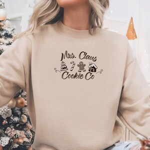 May include: A beige sweatshirt with the text "Mrs. Claus Cookie Co" in a black script. The design includes illustrations of a Christmas tree, candy cane, gingerbread man, and a gingerbread house. The sweatshirt is a crew neck style.