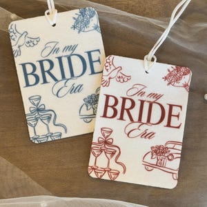 May include: Two white car air fresheners with a floral design and the text "In my Bride Era". One air freshener has blue accents and the other has red accents.