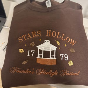 May include: A brown sweatshirt with embroidered text and a gazebo design. The text reads "STARS HOLLOW 1779 Founder's Firelight Festival" in orange thread, with leaf and star accents. The sweatshirt is folded neatly.