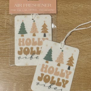 May include: Festive air fresheners with a Christmas theme. The rectangular air fresheners display Christmas trees in green, pink, and brown, with the words "HOLLY JOLLY vibes" in a retro font. The packaging reads "AIR FRESHENER FOR THE CAR, OFFICE, AND BEYOND."
