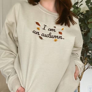 May include: A beige crewneck sweatshirt featuring dark brown embroidered text that reads "I am an autumn." The design is accented with small, colorful embroidered leaves in shades of orange, yellow, and brown scattered around the lettering.