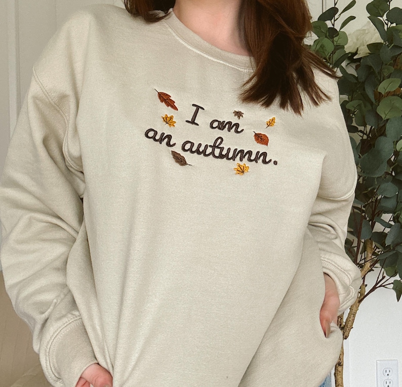 May include: A beige crewneck sweatshirt featuring dark brown embroidered text that reads "I am an autumn." The design is accented with small, colorful embroidered leaves in shades of orange, yellow, and brown scattered around the lettering.