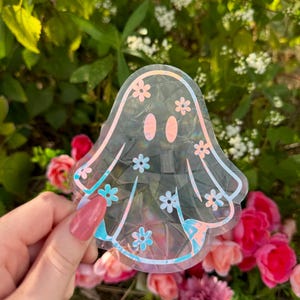 May include: A clear iridescent ghost sticker with a floral pattern. The sticker has a holographic finish and is shaped like a ghost with a rounded head and a flowing body.