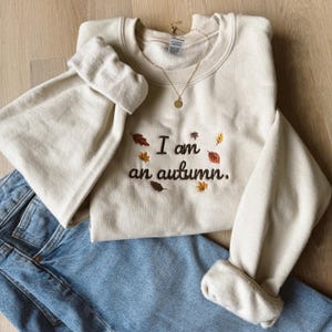 May include: A cream-colored crewneck sweatshirt featuring dark brown embroidered text that reads I am an autumn. The design is accented with small, colorful embroidered leaves in shades of red, orange, and brown, paired with blue denim jeans.