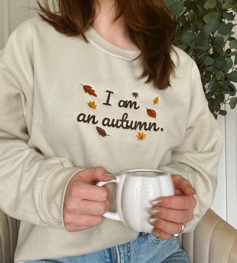 May include: A beige crewneck sweatshirt featuring dark brown embroidered text that reads I am an autumn. The embroidery is accented with small, colorful stitched leaves in shades of orange, yellow, and brown scattered around the lettering.