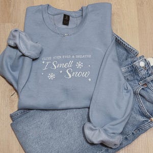 May include: A light blue sweatshirt with the text "Close your eyes & breathe I Smell Snow" in white with snowflake graphics.