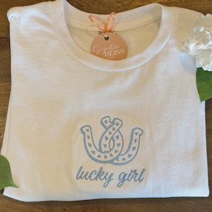 May include: White t-shirt with a blue embroidered design of two horseshoes with hearts inside and the text "lucky girl" underneath.