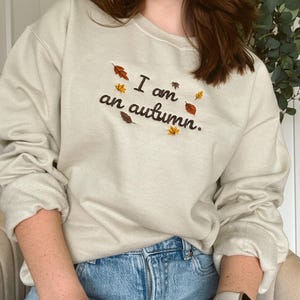 May include: A beige crewneck sweatshirt featuring dark brown embroidered text that reads I am an autumn. The design is accented with small, colorful embroidered leaves in shades of orange, yellow, and brown scattered around the lettering.