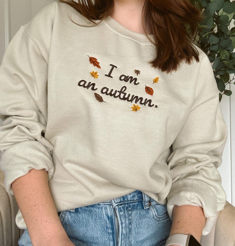 May include: A beige crewneck sweatshirt featuring dark brown embroidered text that reads I am an autumn. The design is accented with small, colorful embroidered leaves in shades of orange, yellow, and brown scattered around the lettering.