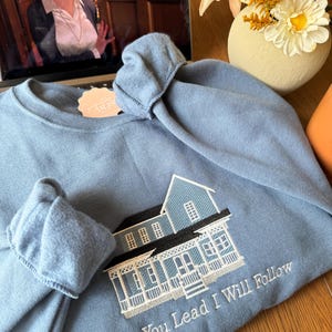 May include: A light blue sweatshirt with an embroidered house design. The house has a blue exterior, black roof, and white trim. The text "Where You Lead I Will Follow" is embroidered below the house. A tag is attached to the sweatshirt.