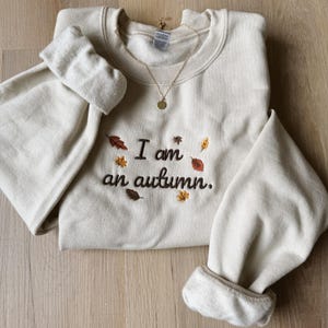 May include: A cream-colored crewneck sweatshirt featuring dark brown embroidered text that reads I am an autumn. The design is accented with small, colorful embroidered leaves in shades of orange, yellow, and brown. A gold-toned necklace rests on the collar.