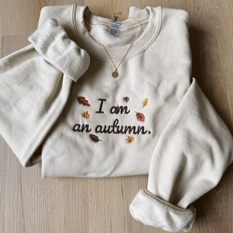 May include: A cream-colored crewneck sweatshirt featuring dark brown embroidered text that reads I am an autumn. The design is accented with small, colorful embroidered leaves in shades of orange, yellow, and brown. A gold-toned necklace rests on the collar.