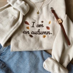 May include: A cream-colored crewneck sweatshirt featuring dark brown embroidered text that reads I am an autumn. The design is accented with small, colorful embroidered fall leaves in shades of red, orange, and yellow, paired with blue jeans.
