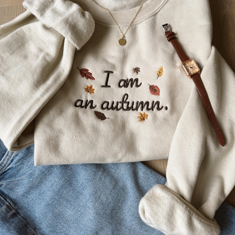 May include: A cream-colored crewneck sweatshirt featuring dark brown embroidered text that reads I am an autumn. The design is accented with small, colorful embroidered fall leaves in shades of red, orange, and yellow, paired with blue jeans.