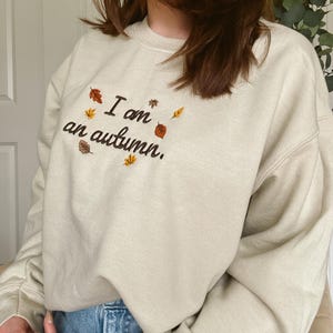 May include: A beige crewneck sweatshirt featuring dark brown embroidered text that reads I am an autumn. The design is accented with small, colorful embroidered leaves in shades of orange, yellow, and brown scattered around the lettering.