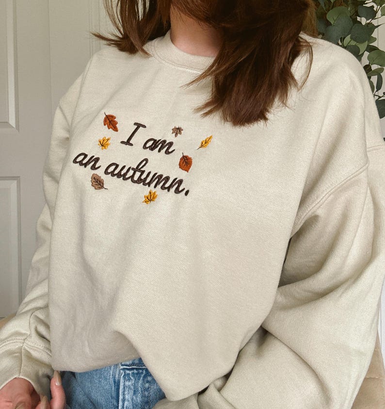 May include: A beige crewneck sweatshirt featuring dark brown embroidered text that reads I am an autumn. The design is accented with small, colorful embroidered leaves in shades of orange, yellow, and brown scattered around the lettering.
