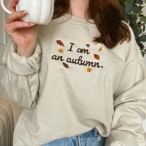 May include: A beige crewneck sweatshirt featuring dark brown embroidered text that reads I am an autumn. The design is accented with small, colorful embroidered leaves in shades of orange, yellow, and brown scattered around the lettering.