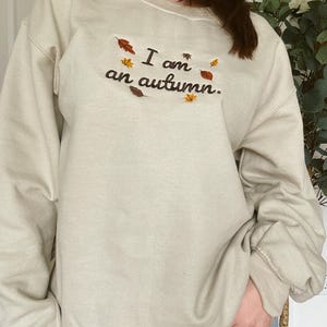 May include: A beige crewneck sweatshirt featuring dark brown embroidered text that reads I am an autumn. The design is accented with small, colorful embroidered leaves in shades of orange, yellow, and brown scattered around the lettering.