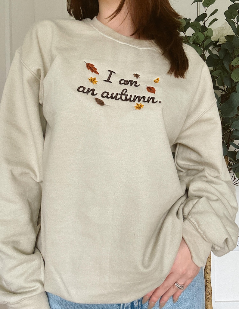 May include: A beige crewneck sweatshirt featuring dark brown embroidered text that reads I am an autumn. The design is accented with small, colorful embroidered leaves in shades of orange, yellow, and brown scattered around the lettering.