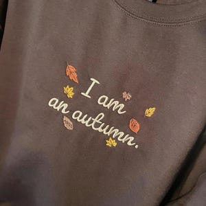 May include: A dark brown crewneck sweatshirt featuring embroidered text that reads "I am an autumn." The embroidery is accented with small, colorful fall leaves in shades of orange, yellow, and brown scattered around the lettering.