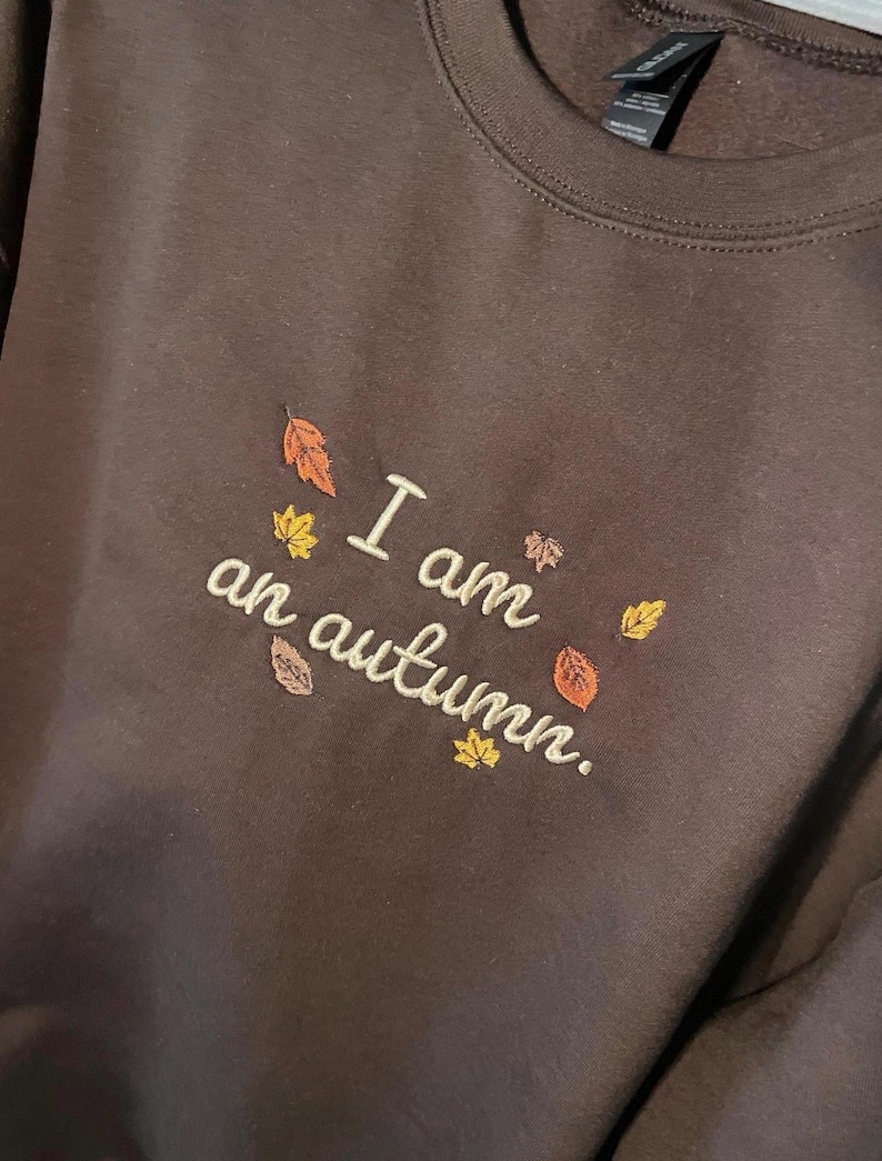 May include: A dark brown crewneck sweatshirt featuring embroidered text that reads "I am an autumn." The embroidery is accented with small, colorful fall leaves in shades of orange, yellow, and brown scattered around the lettering.
