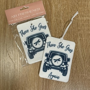 Op de afbeelding: Twee witte autoluchtverfrissers met een blauw Jeep-ontwerp en de tekst "There She Goes" en "There She Goes Again".