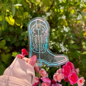 May include: A clear vinyl sticker with a pink and blue outline of a cowboy boot. The boot has a pointed toe and a decorative design on the top.