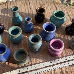 Könnte beinhalten: Eine Sammlung kleiner, handgefertigter Keramikvasen in verschiedenen Farben, darunter Blau, Türkis, Braun und Rosa. Die Vasen sind auf einer Holzoberfläche angeordnet und zeigen ihre einzigartigen Formen und Texturen. Ein Lineal ist im Vordergrund zu sehen.