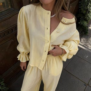 May include: A light yellow two-piece set consisting of a long-sleeved button-down shirt with a round neckline and ruffled cuffs, paired with matching wide-leg trousers. The shirt features large pearl-like buttons along the front placket.