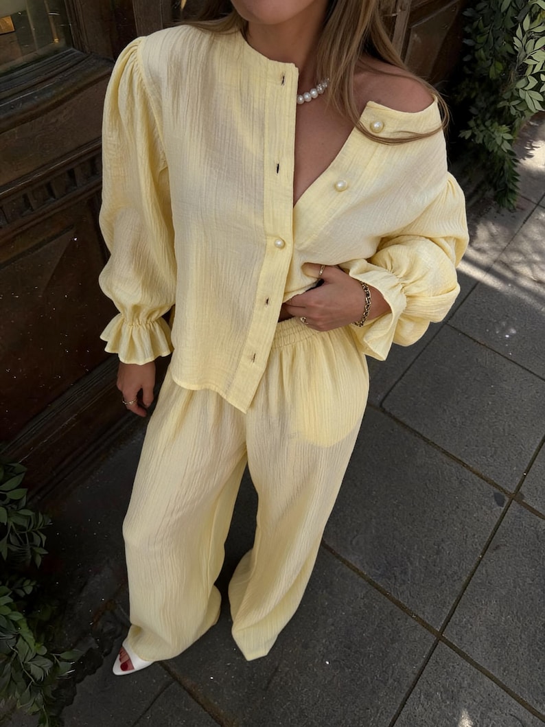 May include: A light yellow two-piece set consisting of a long-sleeved button-down shirt with a round neckline and ruffled cuffs, paired with matching wide-leg trousers. The shirt features large pearl-like buttons along the front placket.
