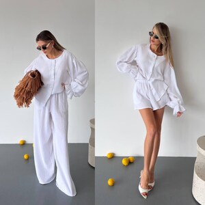 May include: A white two-piece set consisting of a long-sleeved button-down shirt with gathered cuffs and matching wide-leg trousers, or a version with matching shorts. The fabric has a textured, crinkled appearance and a solid white color.