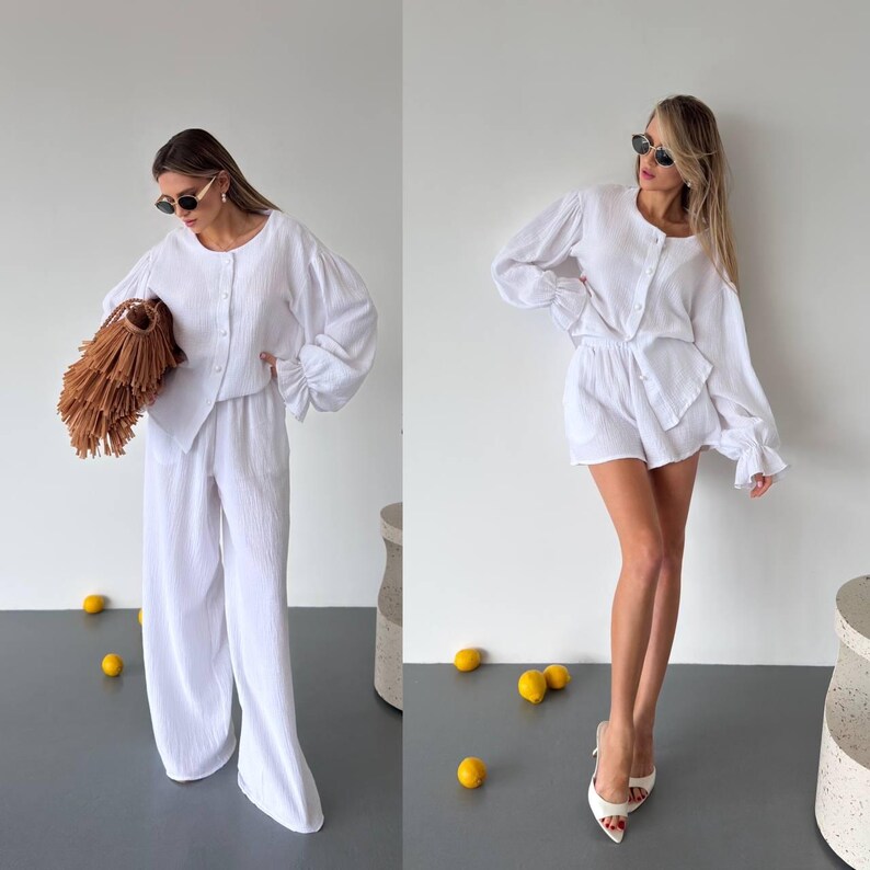 May include: A white two-piece set consisting of a long-sleeved button-down shirt with gathered cuffs and matching wide-leg trousers, or a version with matching shorts. The fabric has a textured, crinkled appearance and a solid white color.