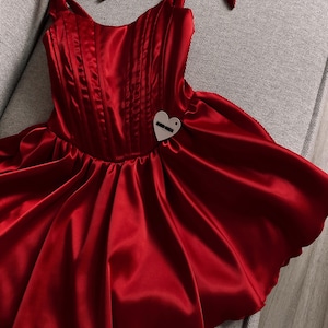 May include: A vibrant red satin dress featuring a structured corset-style bodice with vertical stitching, tie-up shoulder straps, and a flared skirt. A small, heart-shaped white tag with black text is attached to the front of the bodice.