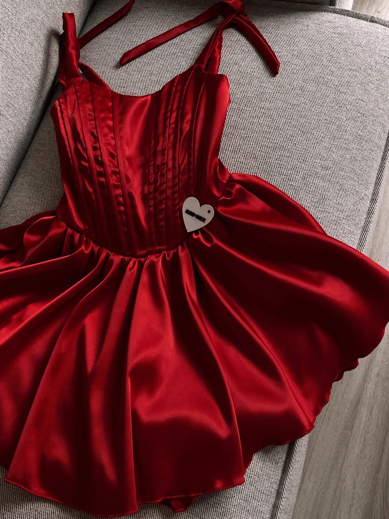 May include: A vibrant red satin dress featuring a structured corset-style bodice with vertical stitching, tie-up shoulder straps, and a flared skirt. A small, heart-shaped white tag with black text is attached to the front of the bodice.