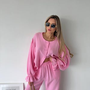 May include: A bright pink two-piece set consisting of a long-sleeved button-down shirt with ruffled cuffs and matching high-waisted trousers. The fabric has a textured, crinkled appearance. The shirt features a round neckline and three buttons.